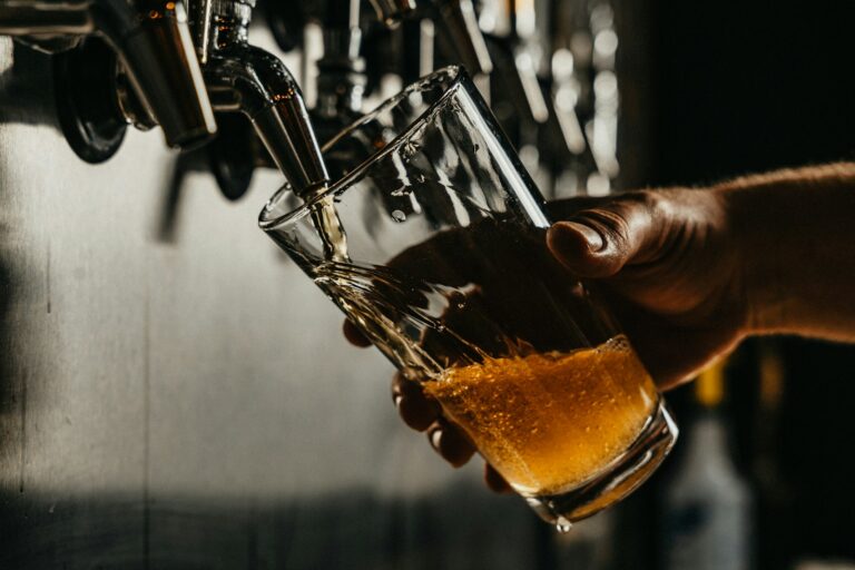 A person's hand holding a pint glass as beer is poured from a tap, with a backdrop of more taps out of focus.